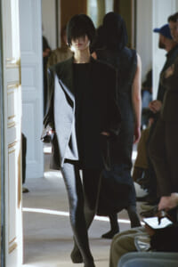 Model in an oversized charcoal suit walking down a fashion show runway, hands in pockets, black outfit underneath.