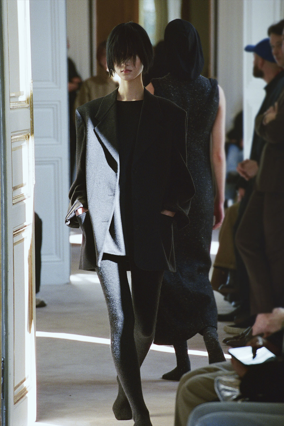 Model in an oversized charcoal suit walking down a fashion show runway, hands in pockets, black outfit underneath.