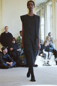 Black female model walking a runway in a dark sleeveless coat over a black dress, carrying a small black handbag; seated audience in the background.