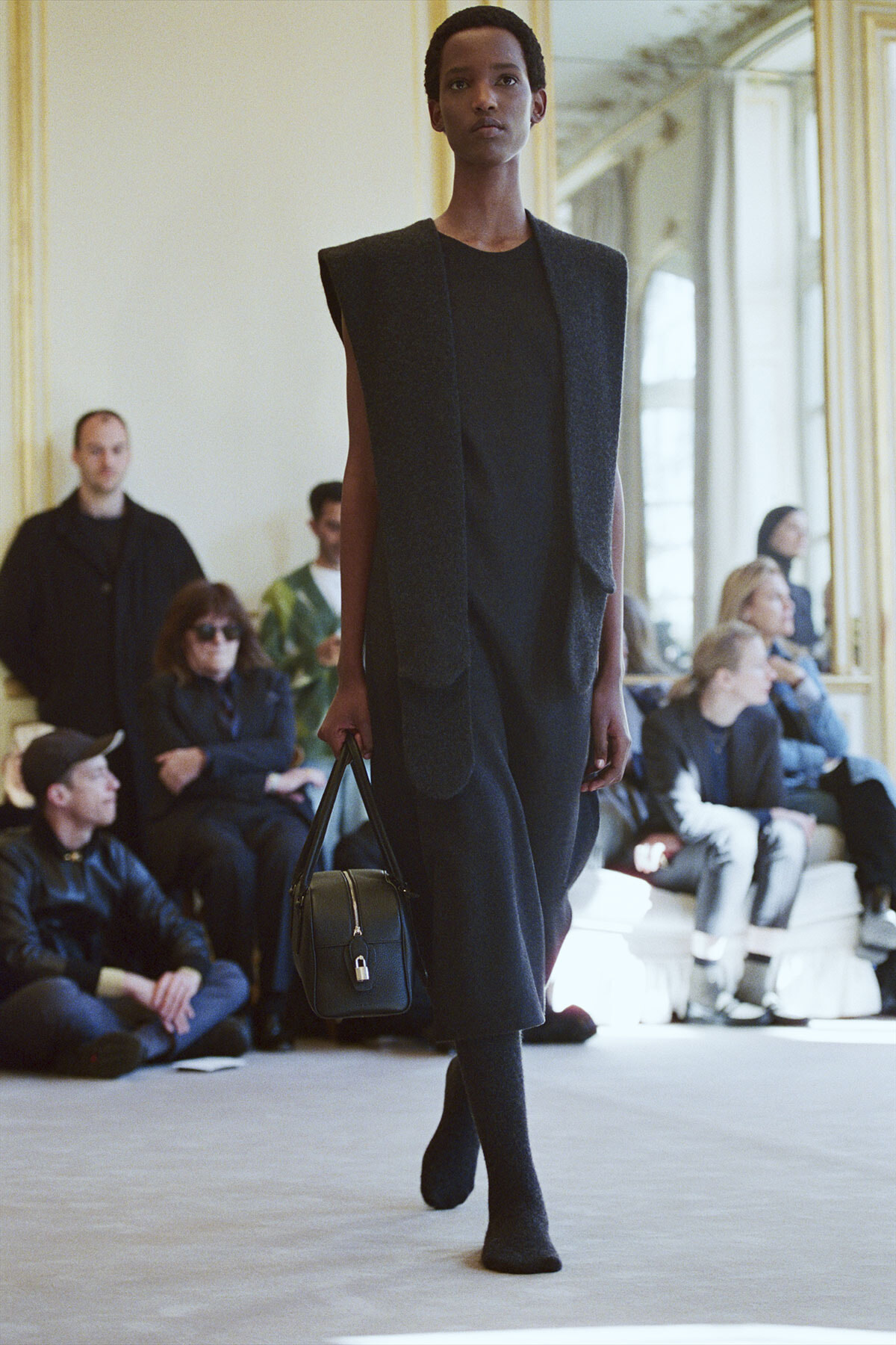 Black female model walking a runway in a dark sleeveless coat over a black dress, carrying a small black handbag; seated audience in the background.