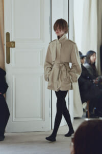 Model wearing a beige belted trench coat and black tights walking past a white door at a fashion show, with seated spectators in the background.