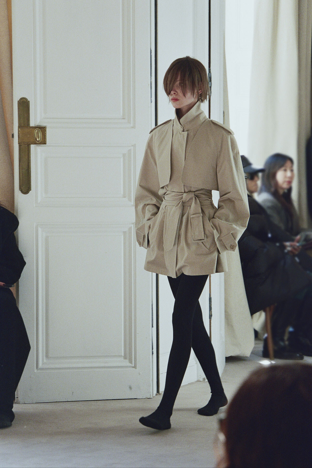 Model wearing a beige belted trench coat and black tights walking past a white door at a fashion show, with seated spectators in the background.