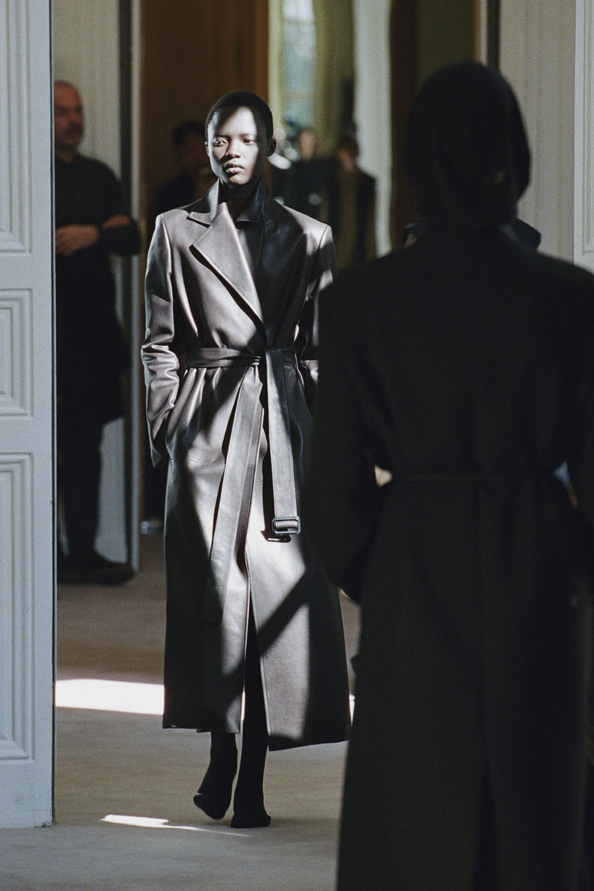 Person wearing a metallic belted trench coat walks through a doorway toward the camera, in a dimly lit hallway.