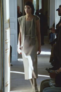 Model in a beige sleeveless dress with a knit scarf walking through a doorway at a fashion show.