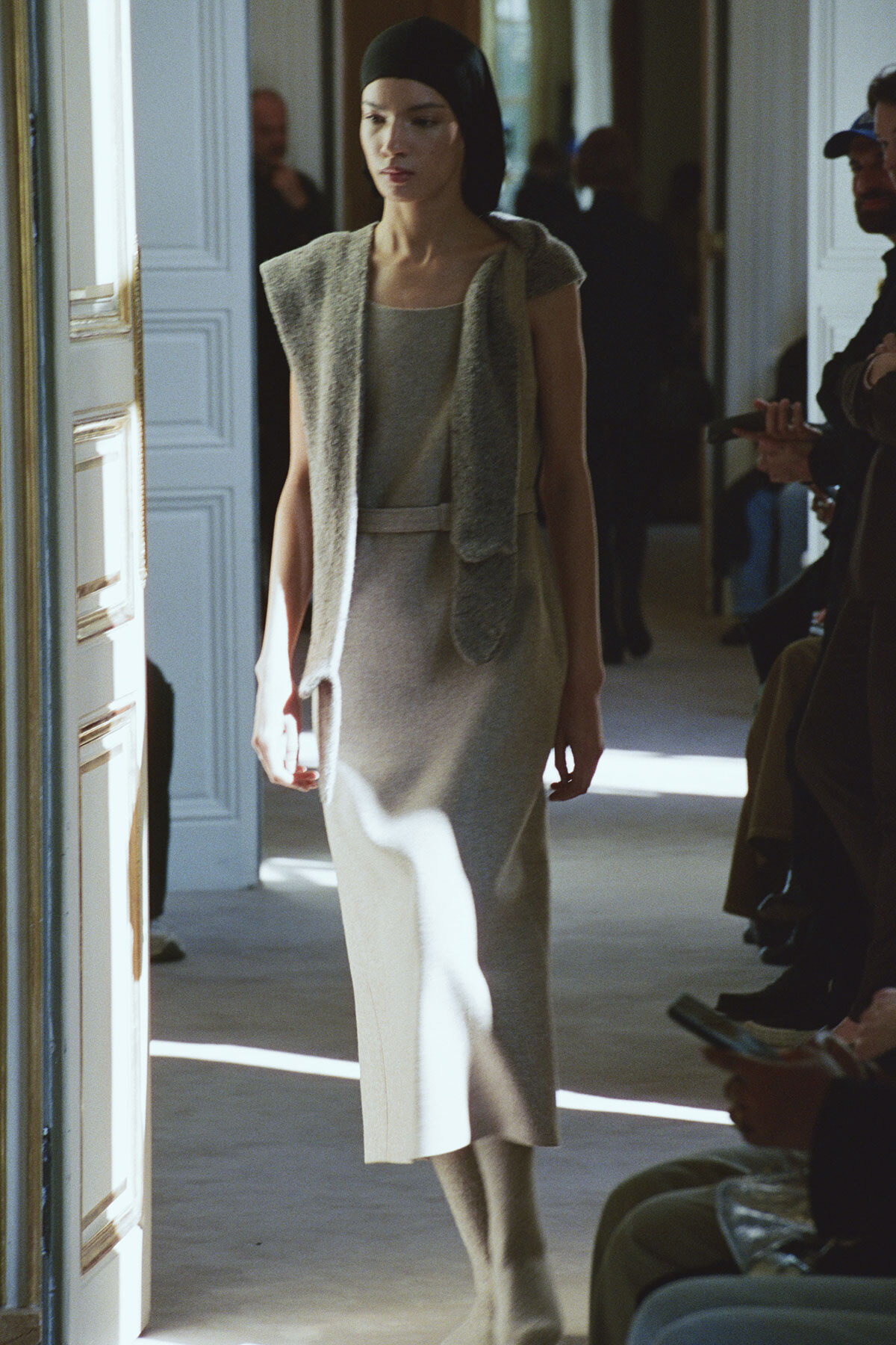 Model in a beige sleeveless dress with a knit scarf walking through a doorway at a fashion show.