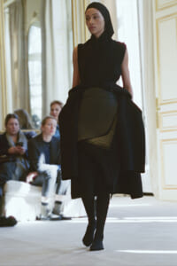 Model walking a runway in a black sleeveless dress with a wrapped belt and headscarf, in an ornate interior.
