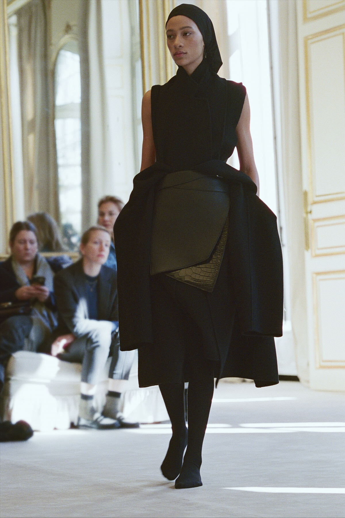Model walking a runway in a black sleeveless dress with a wrapped belt and headscarf, in an ornate interior.