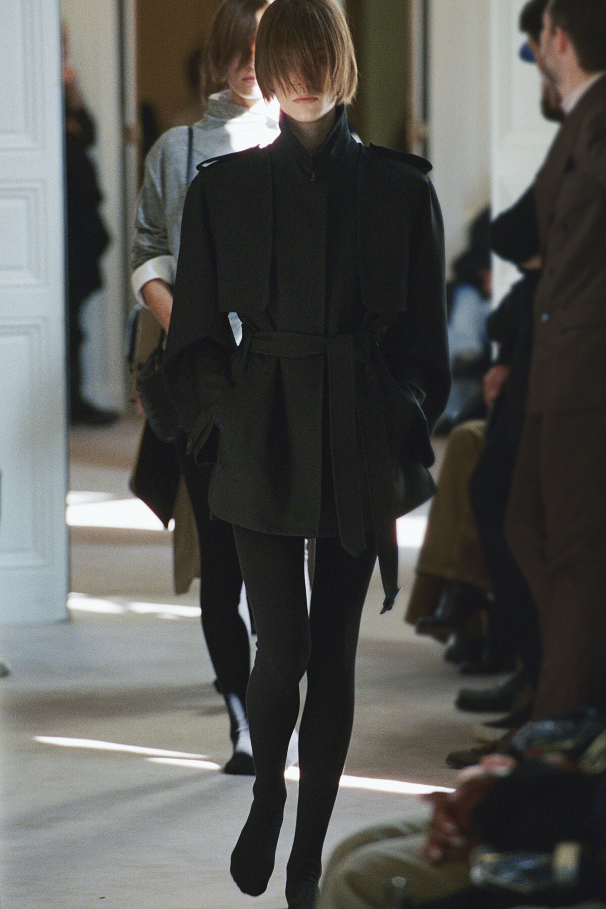 Model walking a fashion runway in a dark belted coat and black tights, with other attendees in the background.