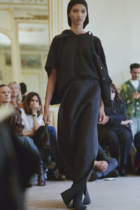 Model walks the runway in a dark gray knit set—oversized top, wide-leg pants, headscarf—carrying a small black bag; audience watches in an ornate room.