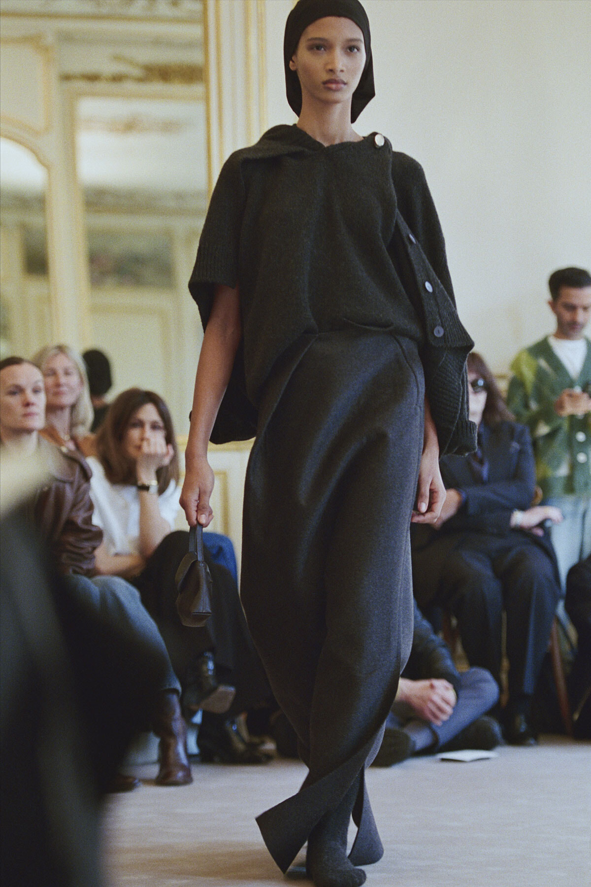 Model walks the runway in a dark gray knit set—oversized top, wide-leg pants, headscarf—carrying a small black bag; audience watches in an ornate room.