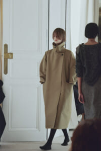 Model wearing a camel-colored, oversized coat with hands in pockets walks past a white door in a fashion setting.