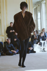 Model walks the runway in a oversized brown fur coat during a fashion show with spectators watching nearby.