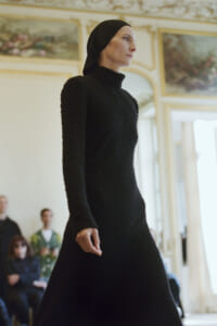 Profile view of a model walking a fashion show in a black textured dress with a headscarf, in an ornate room.
