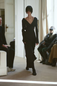 Model walks the runway in a long black off-shoulder dress with slits, indoors at a fashion show.
