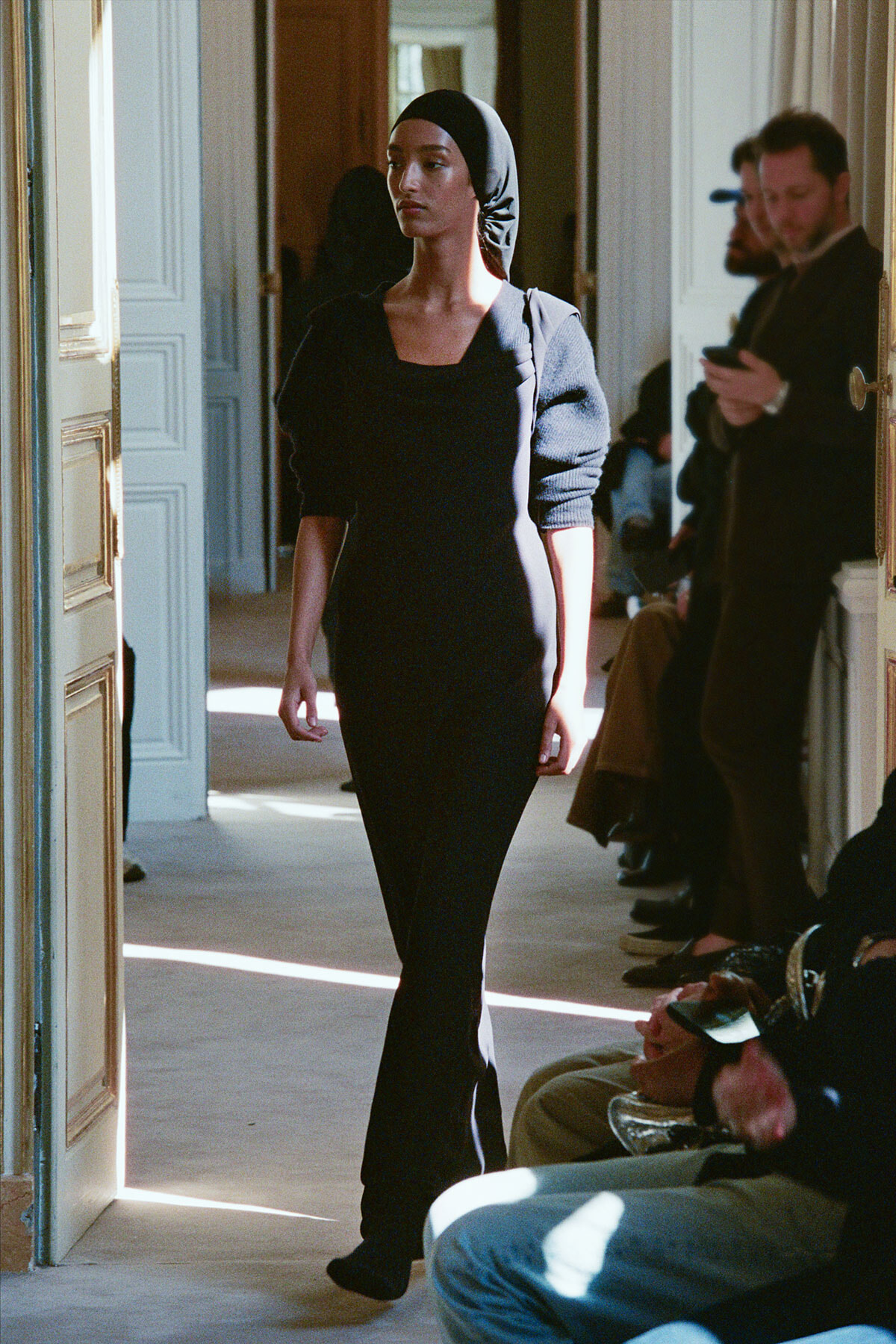 Model walking a fashion show corridor in a black dress and headscarf, with seated spectators along the sides.