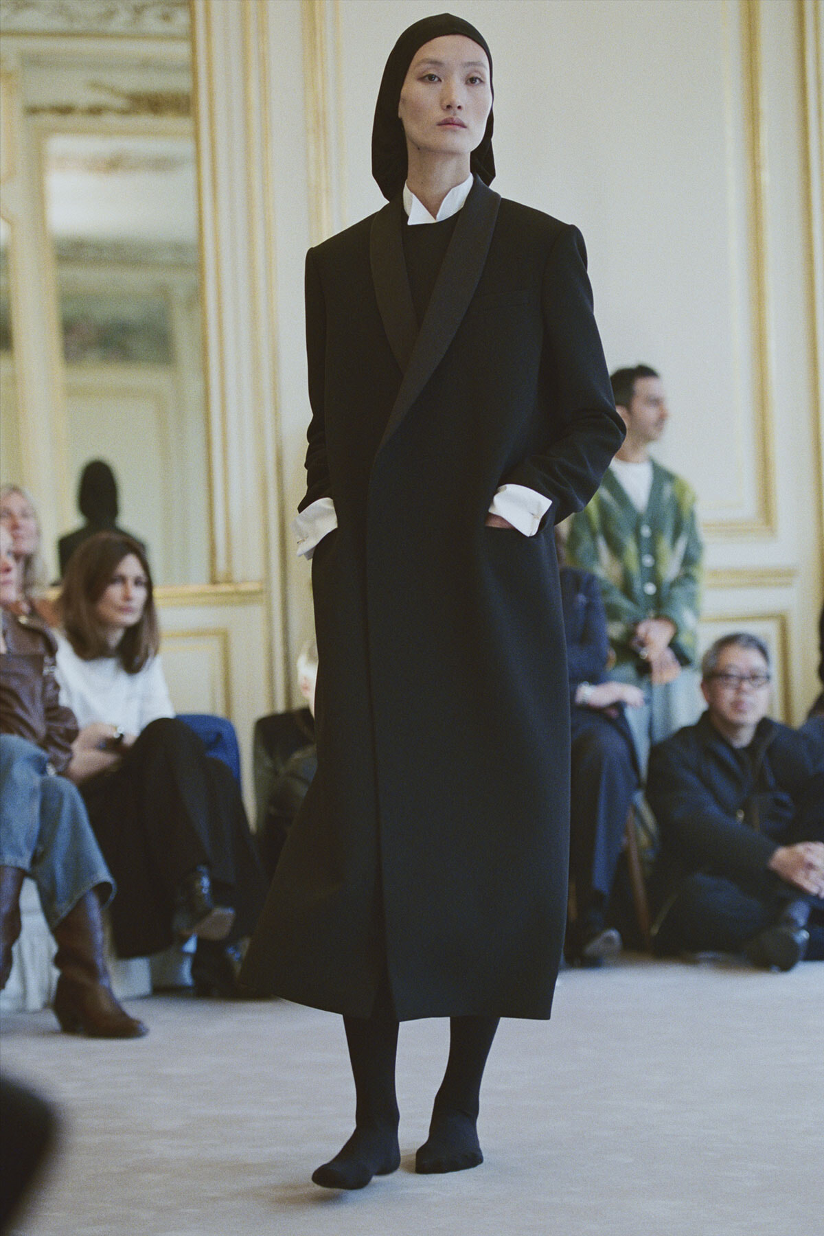 Model walks the runway in a long black coat with wide lapels, white cuffs peeking out, wearing a black headscarf.