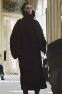 Model wearing a voluminous black coat with a tall, oversized collar stands in a bright, ornate room with mirrors behind.