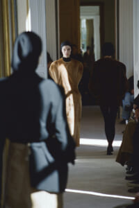 Model walks a fashion show in a tan fur-like coat down a sunlit hallway lined with mirrors; audience seated along the sides.