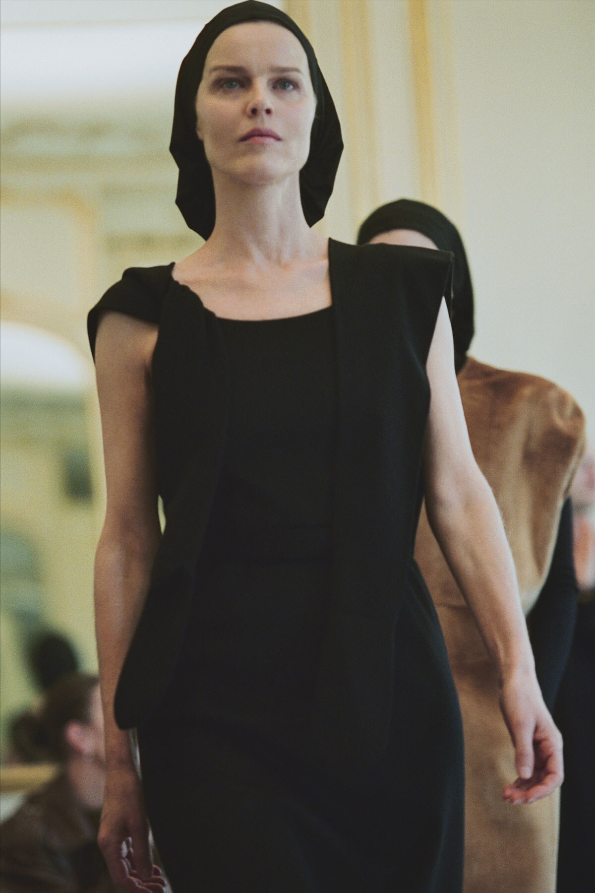 Model walks a runway in a black sleeveless dress with a matching headscarf, background hinting at a fashion show.