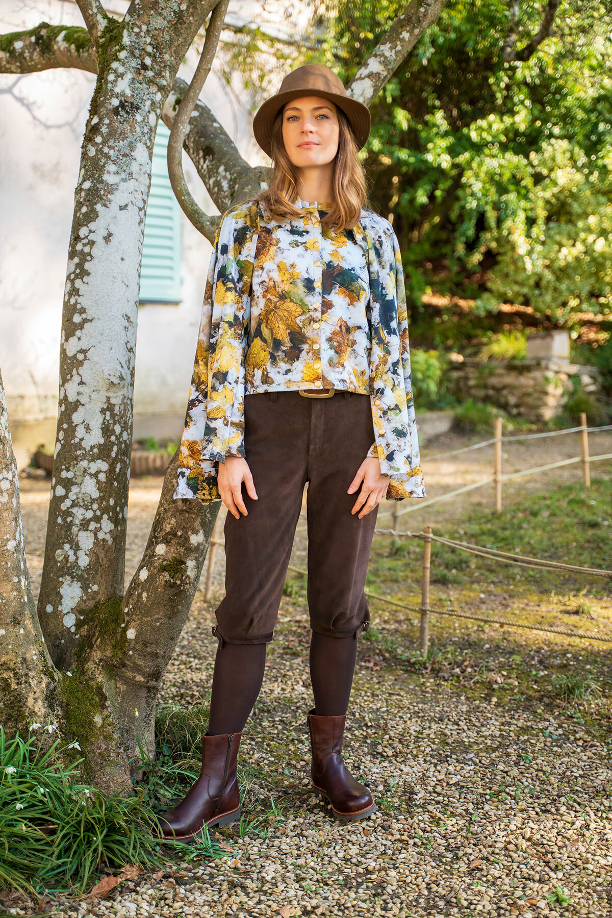 Woman in a wide-brimmed hat standing outdoors beside a tree, wearing a leafy floral blouse, brown pants, and boots.