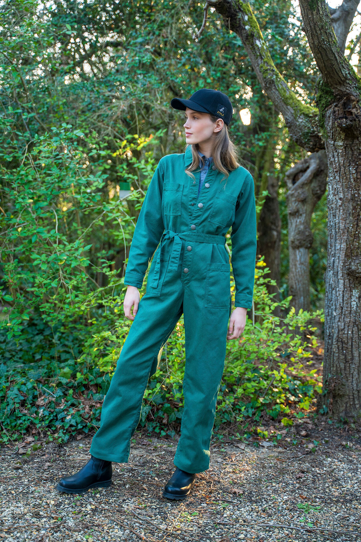 Woman in teal utility jumpsuit with a belt, black cap, and black boots standing in a leafy garden.