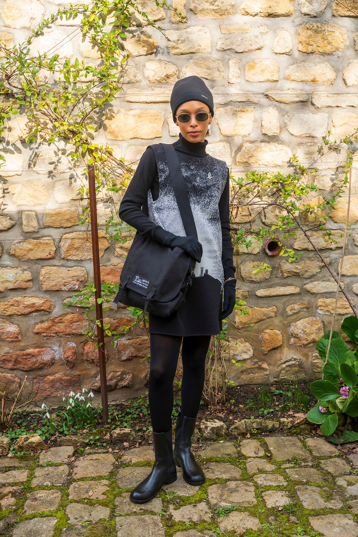 Person in black beanie, sunglasses, and monochrome outfit with a crossbody bag stands in a stone-walled garden.
