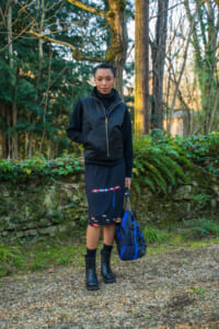 Woman standing on a gravel path in a wooded yard, wearing a black zip vest, black sweater, navy skirt with a red/blue pattern, and holding a blue backpack.