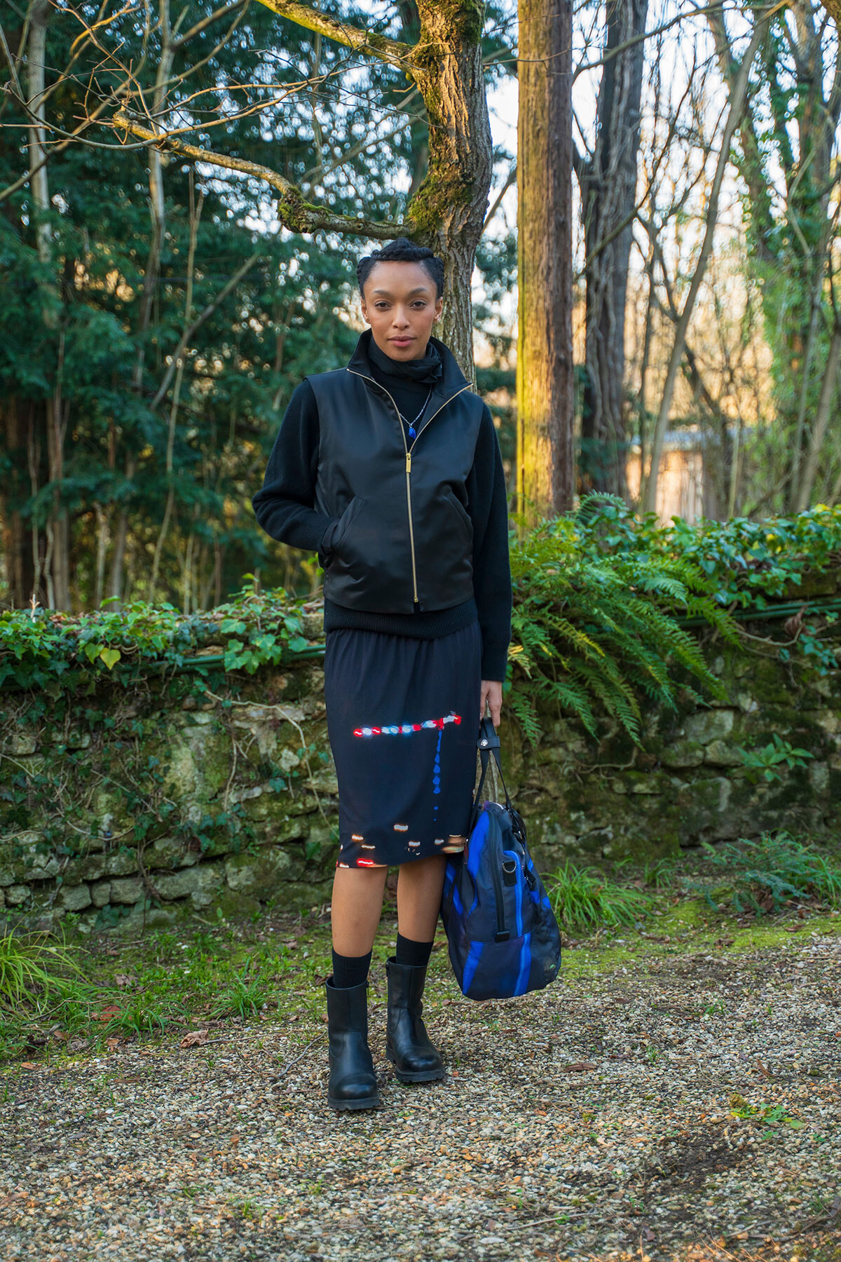 Woman standing on a gravel path in a wooded yard, wearing a black zip vest, black sweater, navy skirt with a red/blue pattern, and holding a blue backpack.