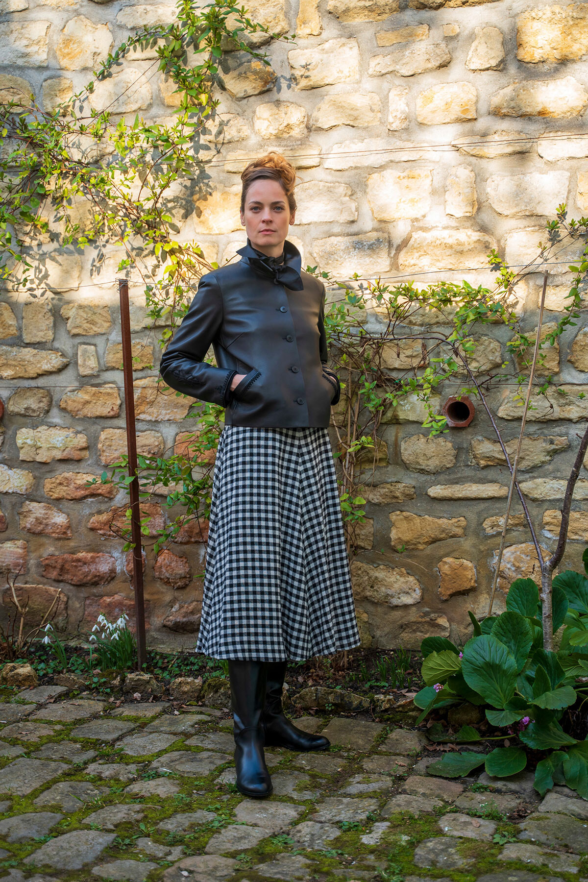 Woman posing in a garden with a stone wall, wearing a black leather jacket, black boots, and a black-and-white gingham midi skirt.