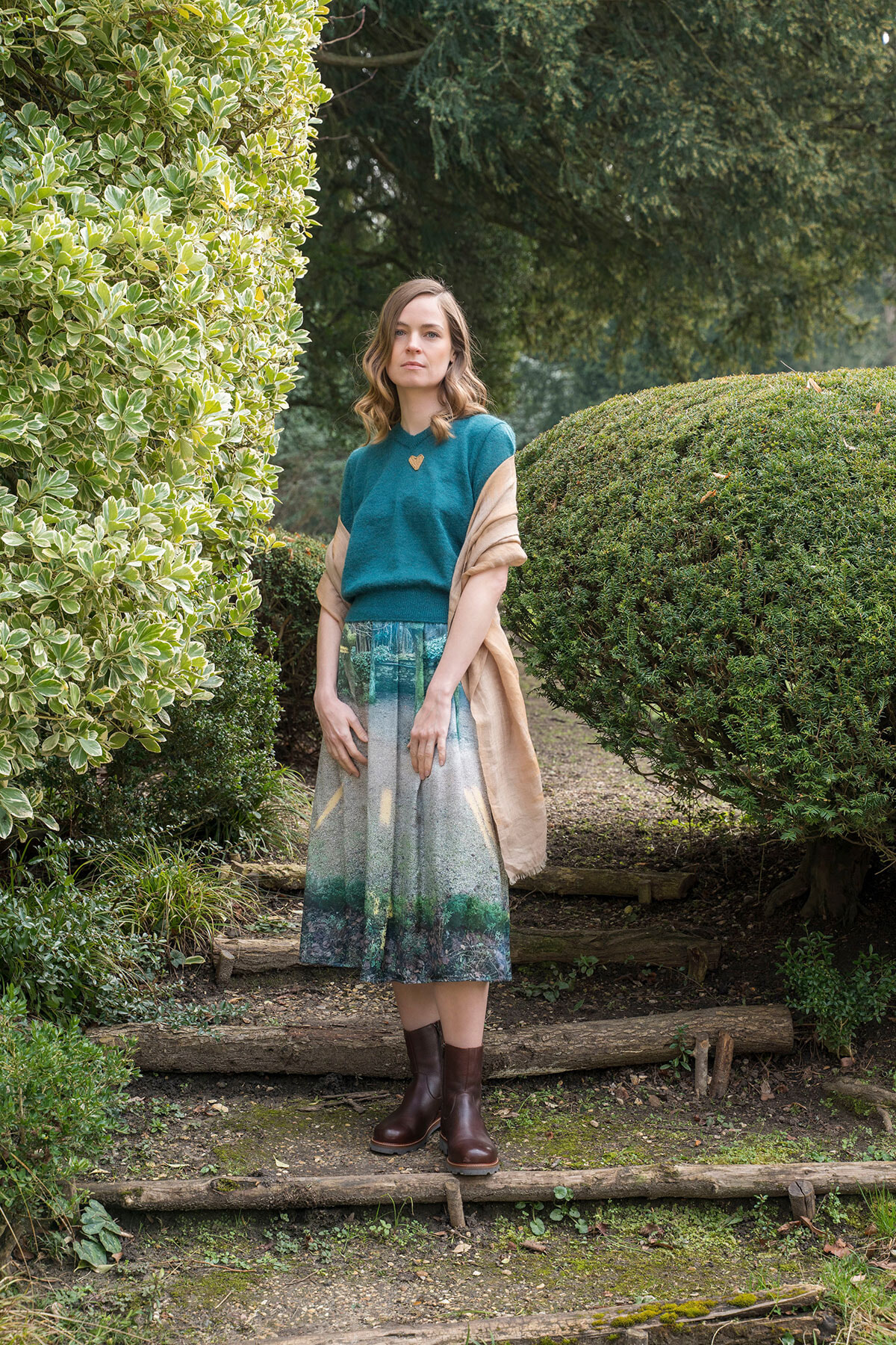 Woman standing on garden steps wearing a teal top, beige shawl, and a printed midi skirt with brown boots.