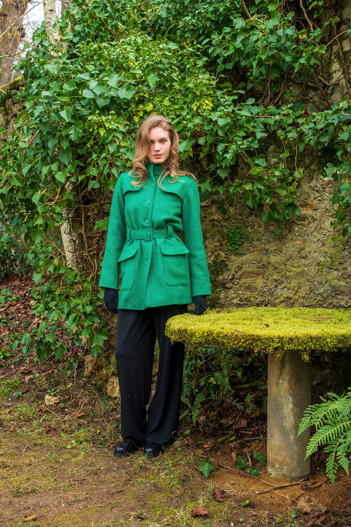 Woman in a bright green belted coat standing outdoors beside a mossy stone table with ivy-covered wall behind her.