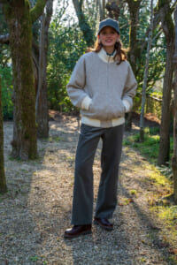 Woman standing on a sunlit forest path wearing a gray bomber jacket, white turtleneck, dark pants, and boots with a cap.