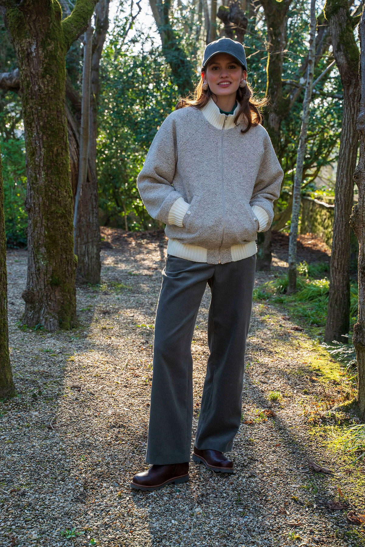 Woman standing on a sunlit forest path wearing a gray bomber jacket, white turtleneck, dark pants, and boots with a cap.