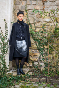 Woman in a black coat and scarf with a monochrome printed skirt standing against an ivy-covered stone wall.