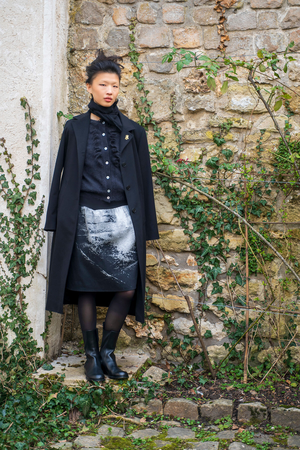 Woman in a black coat and scarf with a monochrome printed skirt standing against an ivy-covered stone wall.