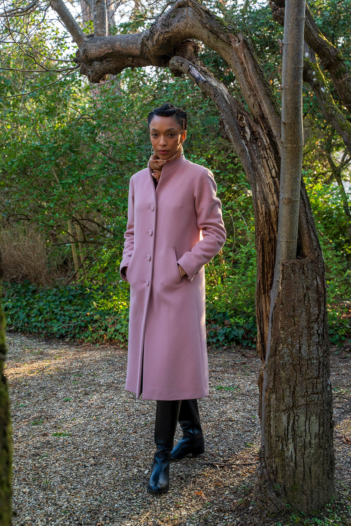 Woman in a pale pink knee-length coat and black boots stands in a garden, posing beside a gnarled tree branch.
