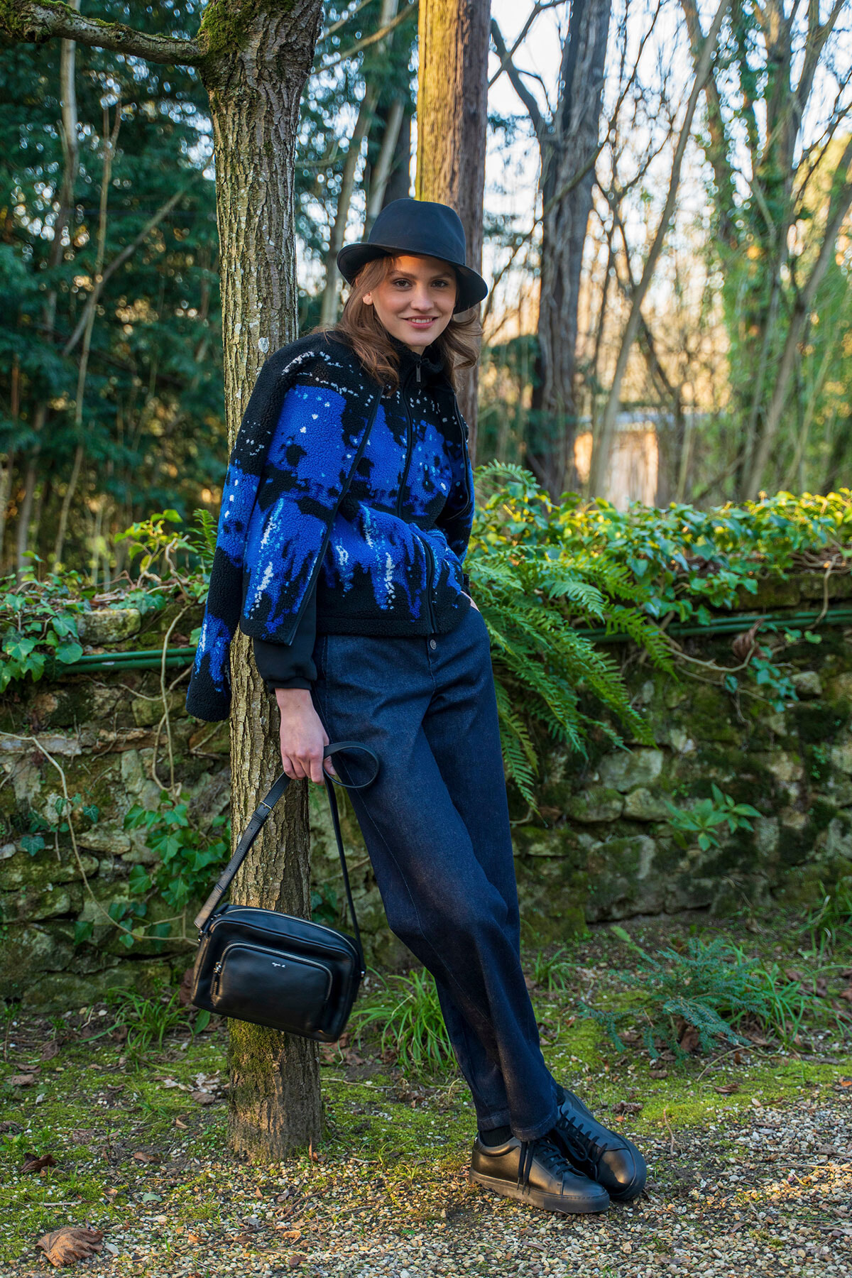 Woman leaning against a tree in a forest, wearing a blue patterned sweater, black hat, dark jeans, and carrying a small black bag.