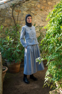Woman in a pale blue, buttoned jacket and matching skirt wearing a black hijab stands in a garden beside a stone wall.