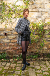 Woman standing against a stone wall in a black-and-white checkered shirt over a black top, leather shorts, tights, and black boots.”