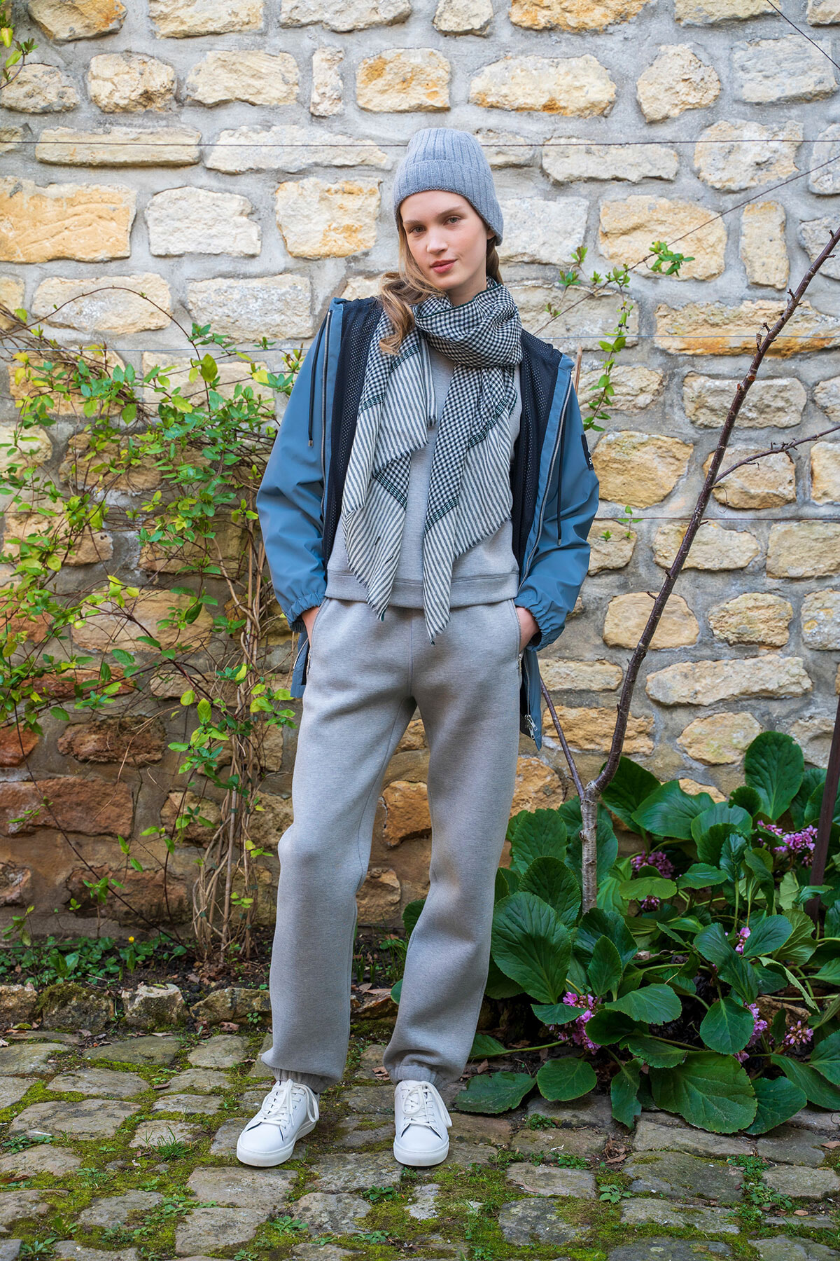 Young woman in a blue jacket, gray beanie, striped scarf, gray sweatpants, and white sneakers posing by a stone wall.