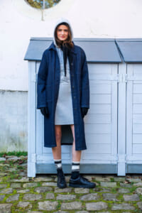 Person standing outdoors in a long blue denim coat over a gray hoodie, black gloves, and sneakers, with a pale blue shed in the background.