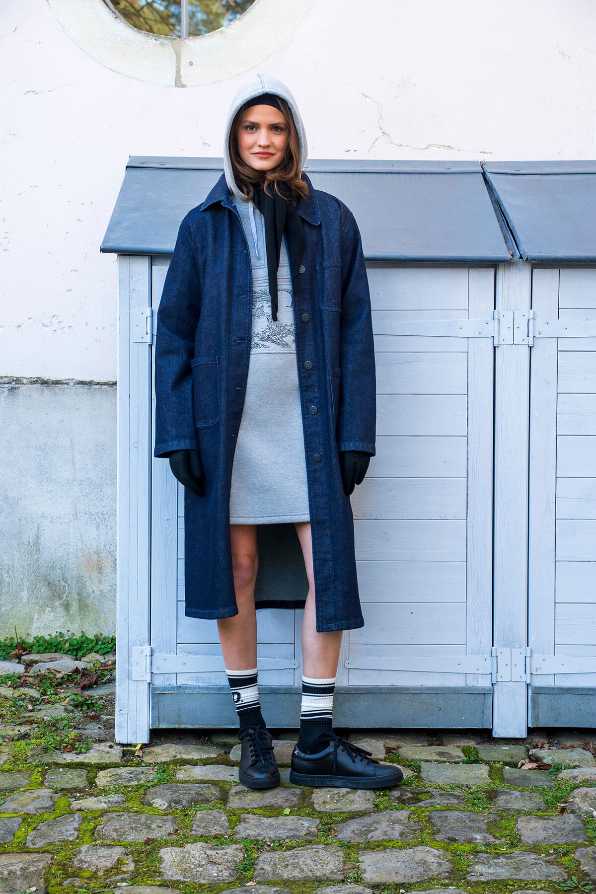 Person standing outdoors in a long blue denim coat over a gray hoodie, black gloves, and sneakers, with a pale blue shed in the background.