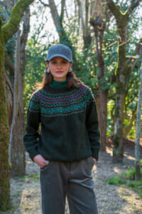 Woman standing in a forest wearing a dark green knit sweater with a colorful yoke pattern, gray cap, and corduroy pants, hands in pockets.