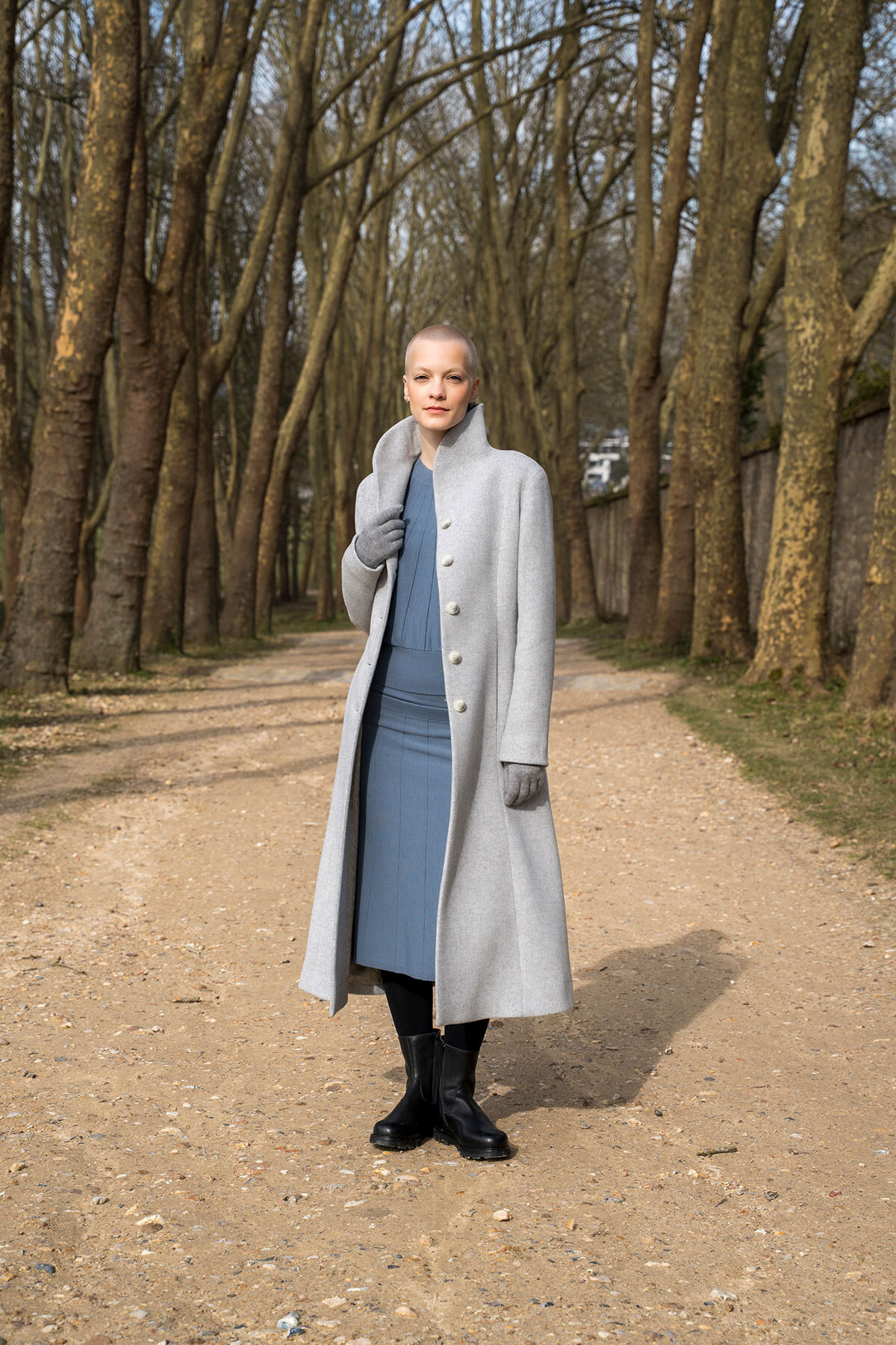 Person standing on a tree-lined path wearing a long light gray coat, blue dress, gloves, and black boots.