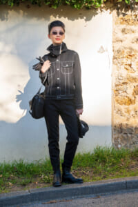 Person wearing an all-black outfit (denim jacket, pants, boots) with sunglasses, holding gloves and a bag outdoors by a white wall.
