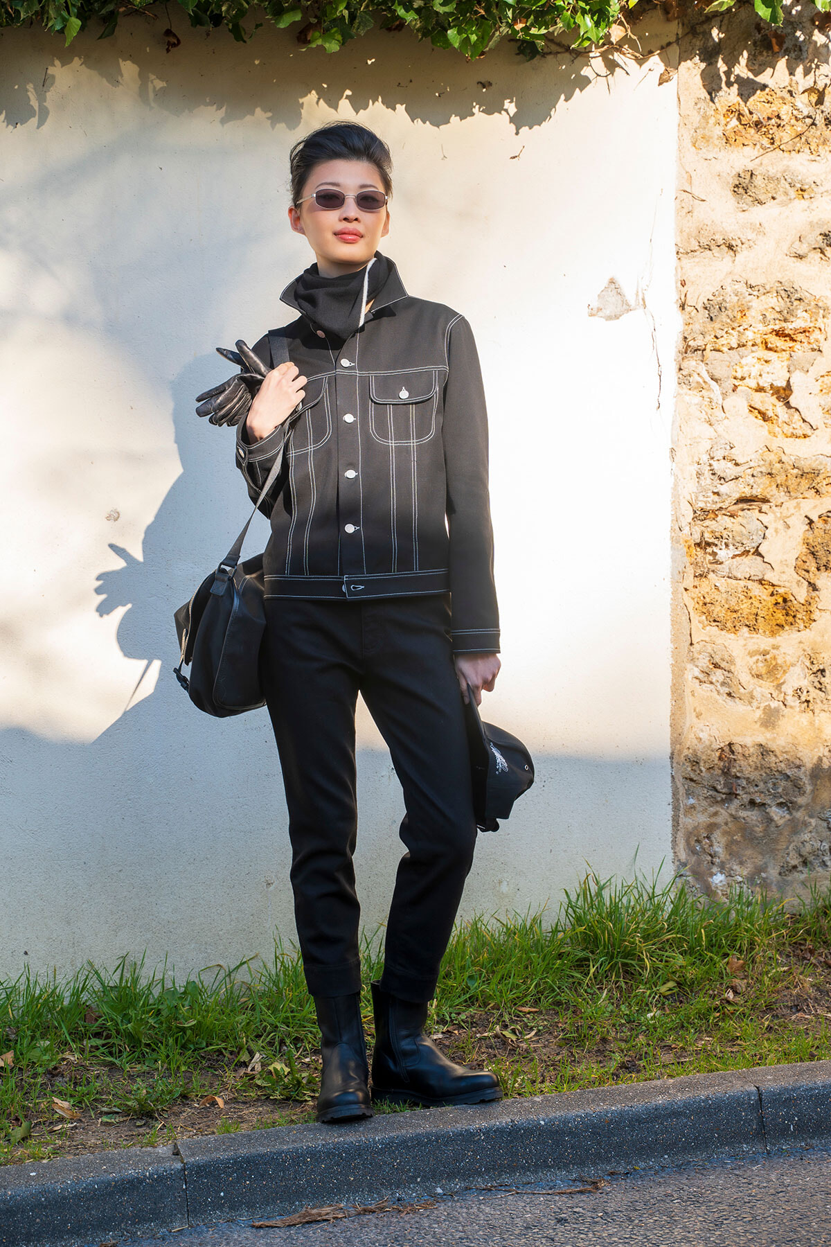 Person wearing an all-black outfit (denim jacket, pants, boots) with sunglasses, holding gloves and a bag outdoors by a white wall.