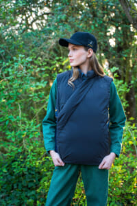 Woman posed outdoors in a navy puffer vest layered over a green tracksuit, wearing a black cap with a logo, looking to the side.