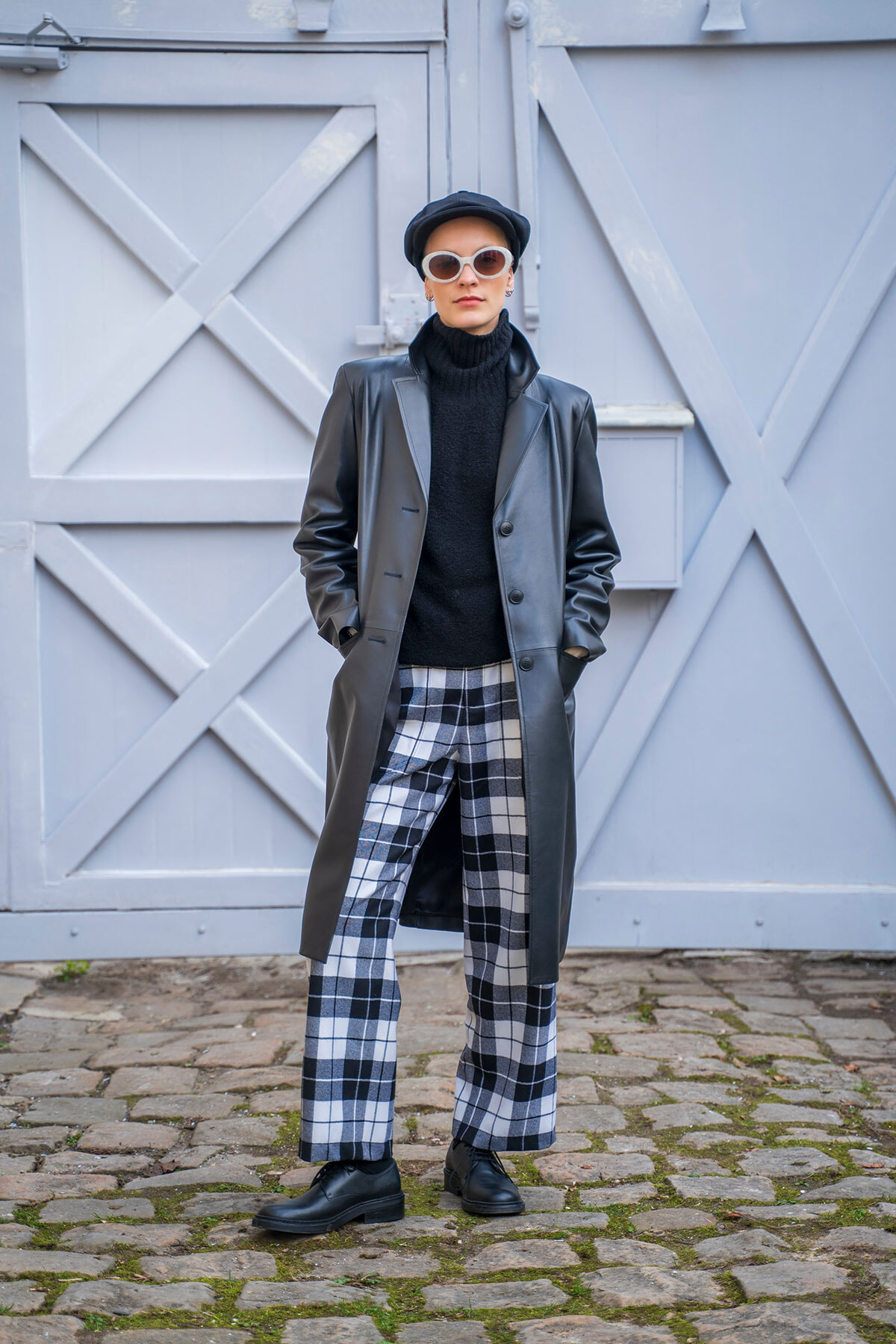 Stylish person in a black turtleneck and long gray leather coat, plaid pants, and white sunglasses, posing against pale blue doors on a cobblestone street.