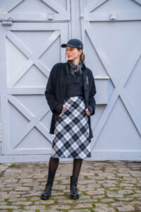 Woman in a navy cap, black coat, and plaid midi skirt posing against pale gray wooden doors in a cobblestone setting.