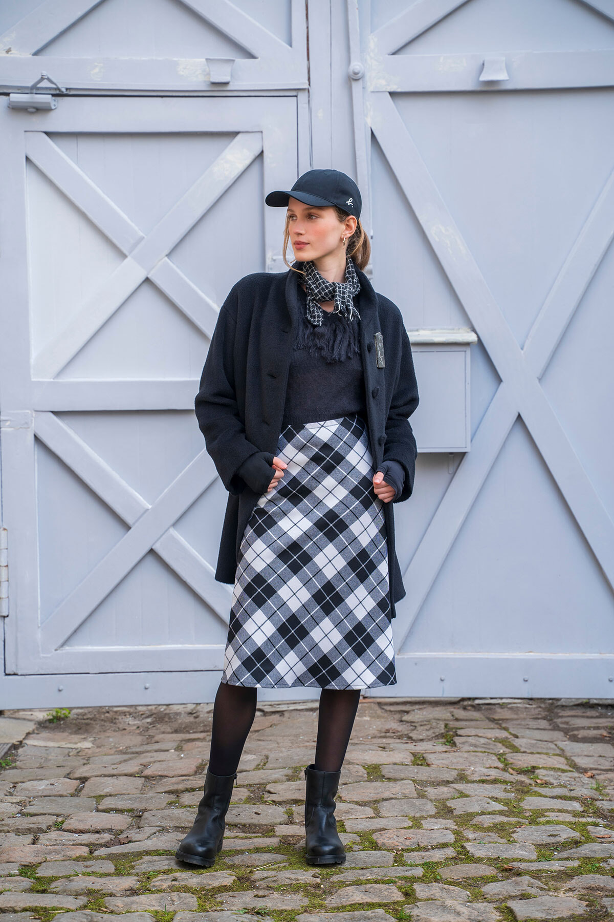 Woman in a navy cap, black coat, and plaid midi skirt posing against pale gray wooden doors in a cobblestone setting.